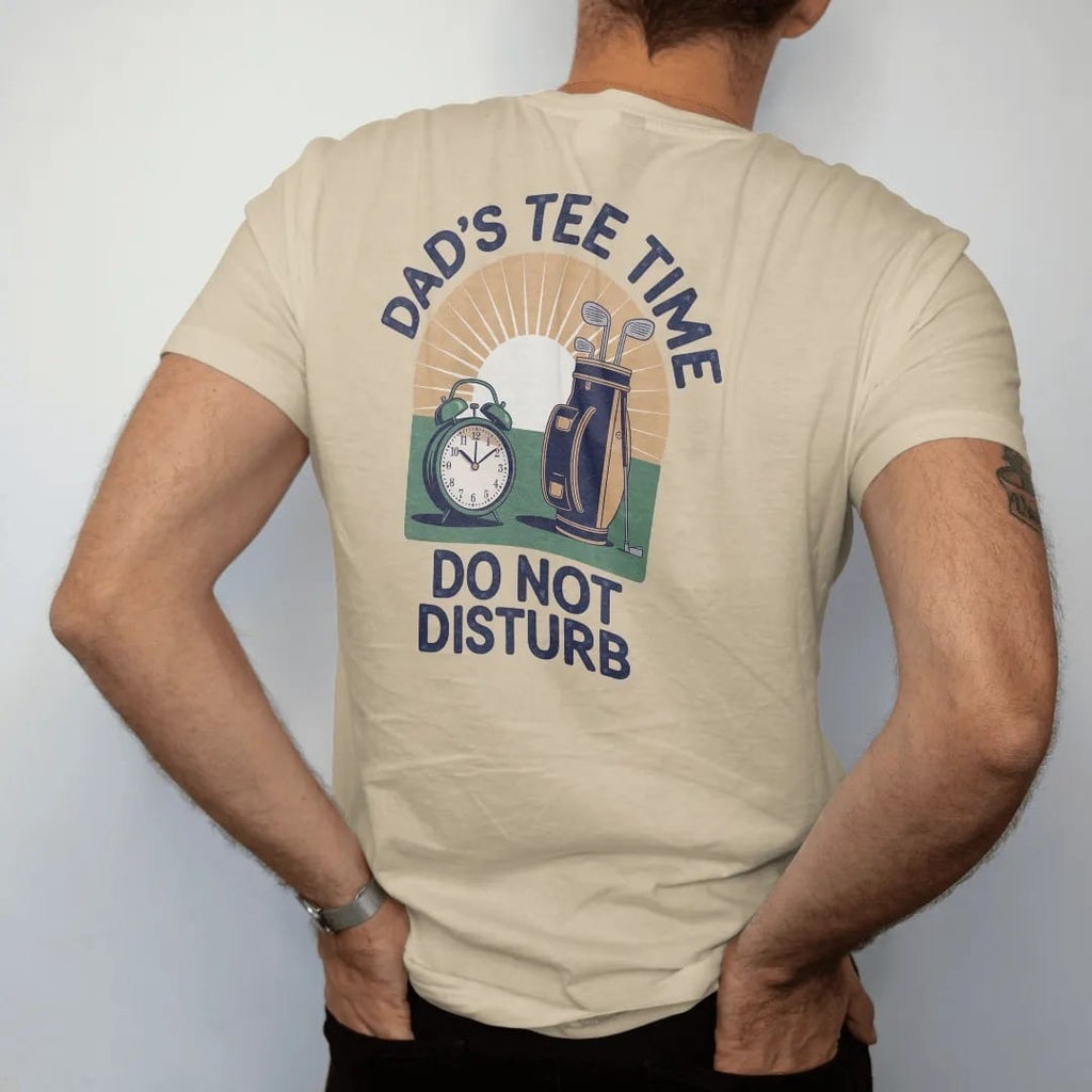 Man wearing a Sandstone tee, seen from the back: a vintage alarm clock beside a golf bag at sunrise, with "DAD'S TEE TIME" and "DO NOT DISTURB" in chunky rounded 70s serif letters, printed in faded fairway green, washed navy, faded flag red and…