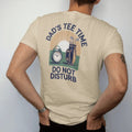 Man wearing a Sandstone tee, seen from the back: a vintage alarm clock beside a golf bag at sunrise, with "DAD'S TEE TIME" and "DO NOT DISTURB" in chunky rounded 70s serif letters, printed in faded fairway green, washed navy, faded flag red and…