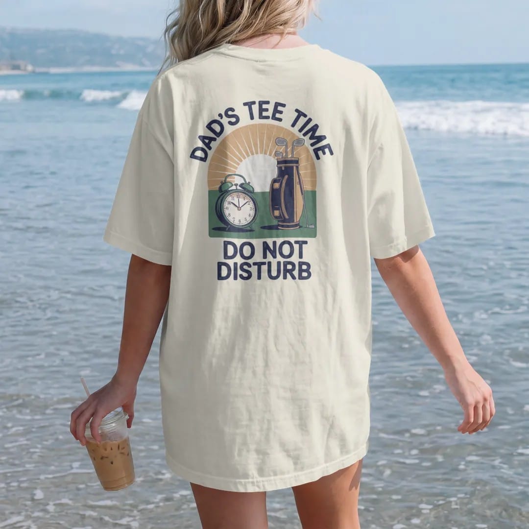 Woman on the beach in a Ivory tee, back view: a vintage alarm clock beside a golf bag at sunrise, with "DAD'S TEE TIME" and "DO NOT DISTURB" in chunky rounded 70s serif letters, printed in faded fairway green, washed navy, faded flag red and sand…