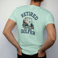 Man wearing a Chalky Mint tee, seen from the back: an older golfer in a bucket hat waving from a golf cart, with "RETIRED" and "FULL-TIME GOLFER" in loopy 1970s script, printed in faded fairway green, washed navy, faded flag red and sand tan with a…