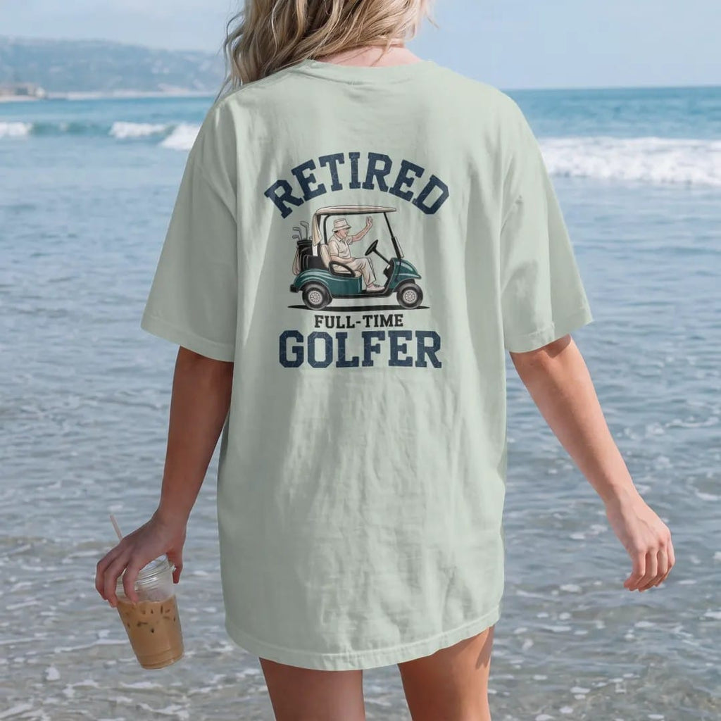 Woman on the beach in a Bay tee, back view: an older golfer in a bucket hat waving from a golf cart, with "RETIRED" and "FULL-TIME GOLFER" in loopy 1970s script, printed in faded fairway green, washed navy, faded flag red and sand tan with a…