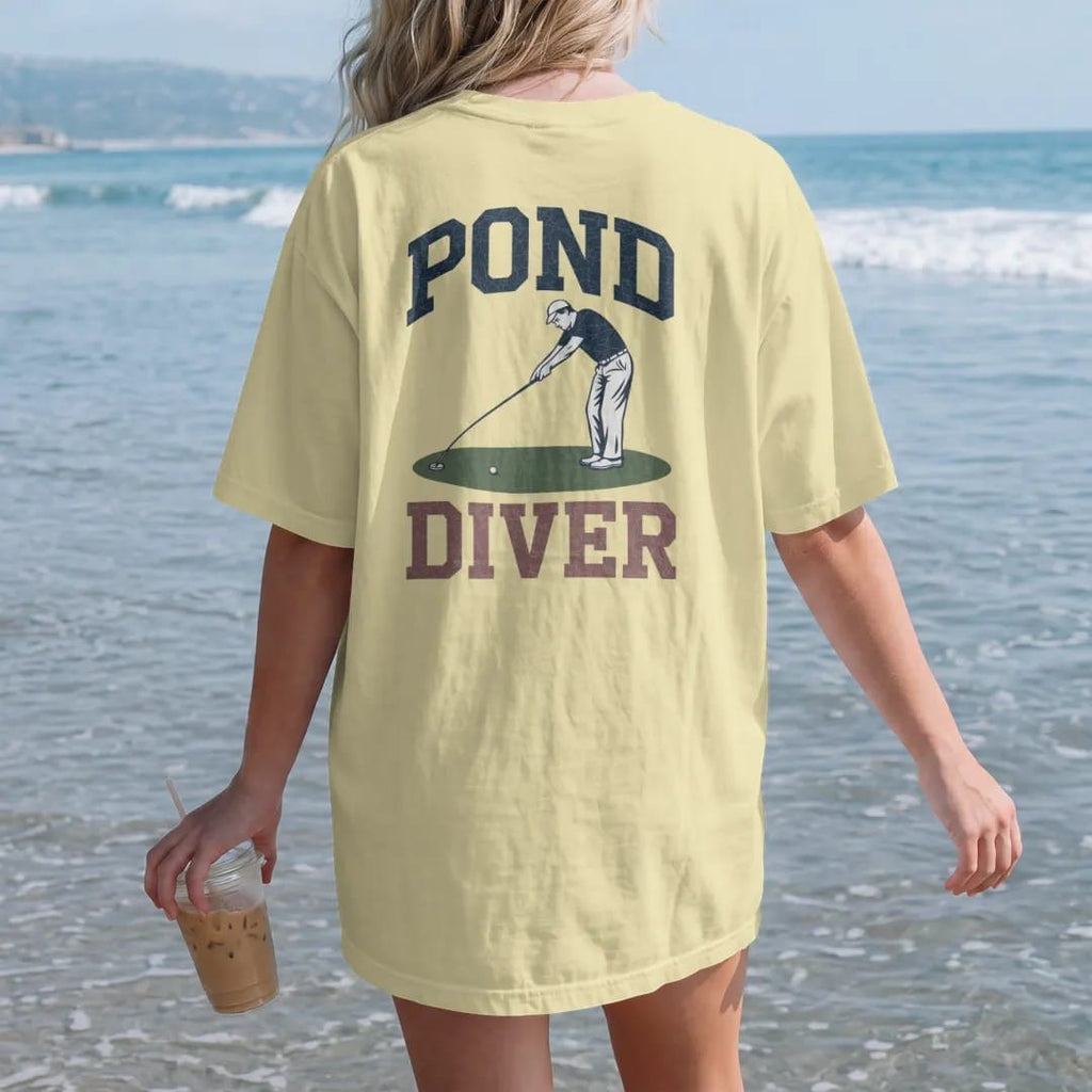 Woman on the beach in a Butter tee, back view: a golfer stretching a long ball-retriever pole to fish a ball out of a pond, with "POND" and "DIVER" in chunky faded collegiate block letters, printed in faded fairway green, washed navy, faded flag…