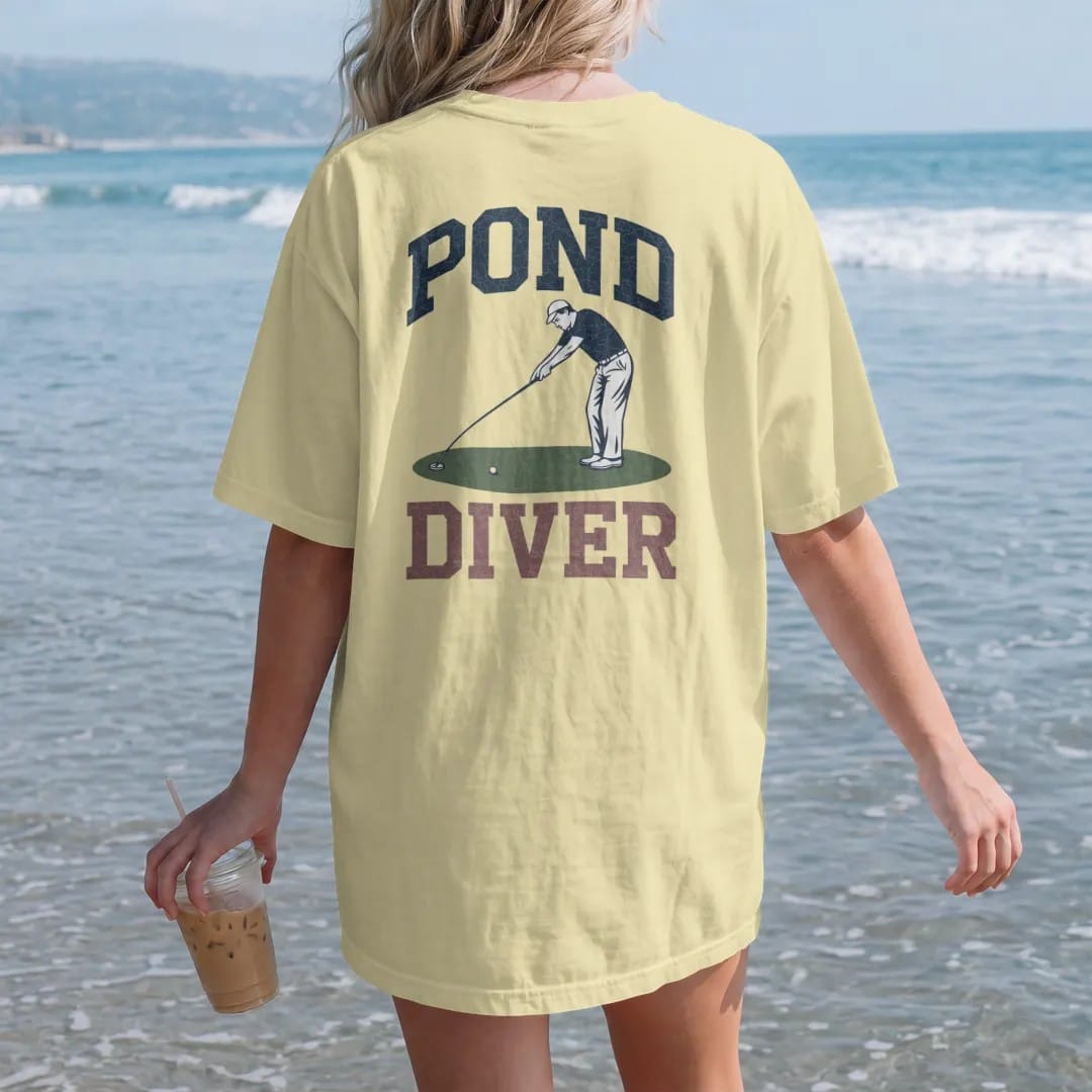 Woman on the beach in a Butter tee, back view: a golfer stretching a long ball-retriever pole to fish a ball out of a pond, with "POND" and "DIVER" in chunky faded collegiate block letters, printed in faded fairway green, washed navy, faded flag…