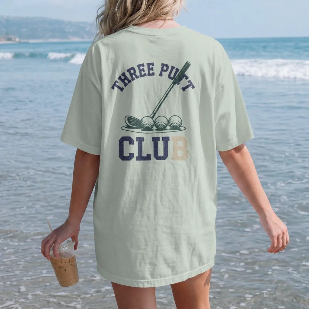 Woman on the beach in a Bay tee, back view: a putter and three golf balls in a row near the cup, with "THREE PUTT" and "CLUB" in chunky faded collegiate block letters, printed in faded fairway green, washed navy, faded flag red and sand tan with a…