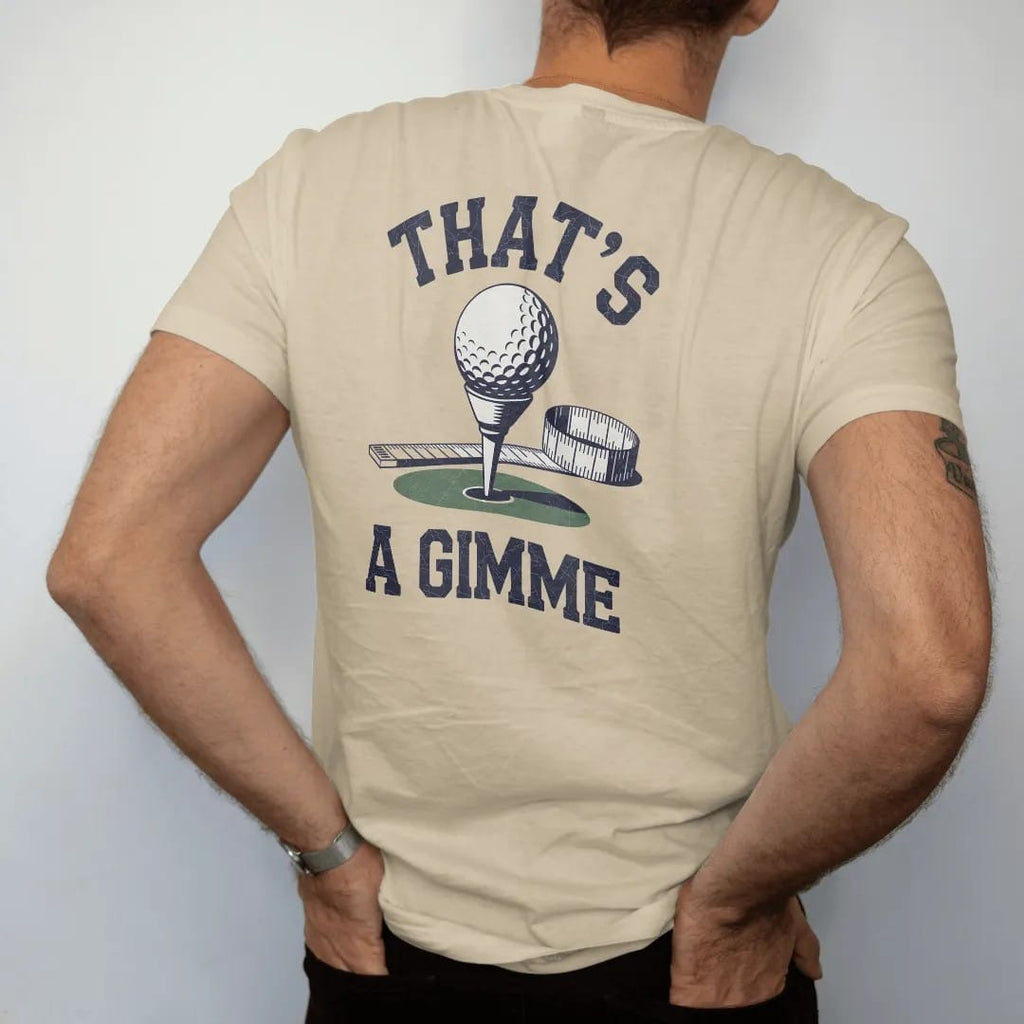 Man wearing a Sandstone tee, seen from the back: a golf ball resting a few inches from the cup with a measuring tape stretched beside it, with "THAT'S" and "A GIMME" in loopy 1970s script, printed in faded fairway green, washed navy, faded flag red…