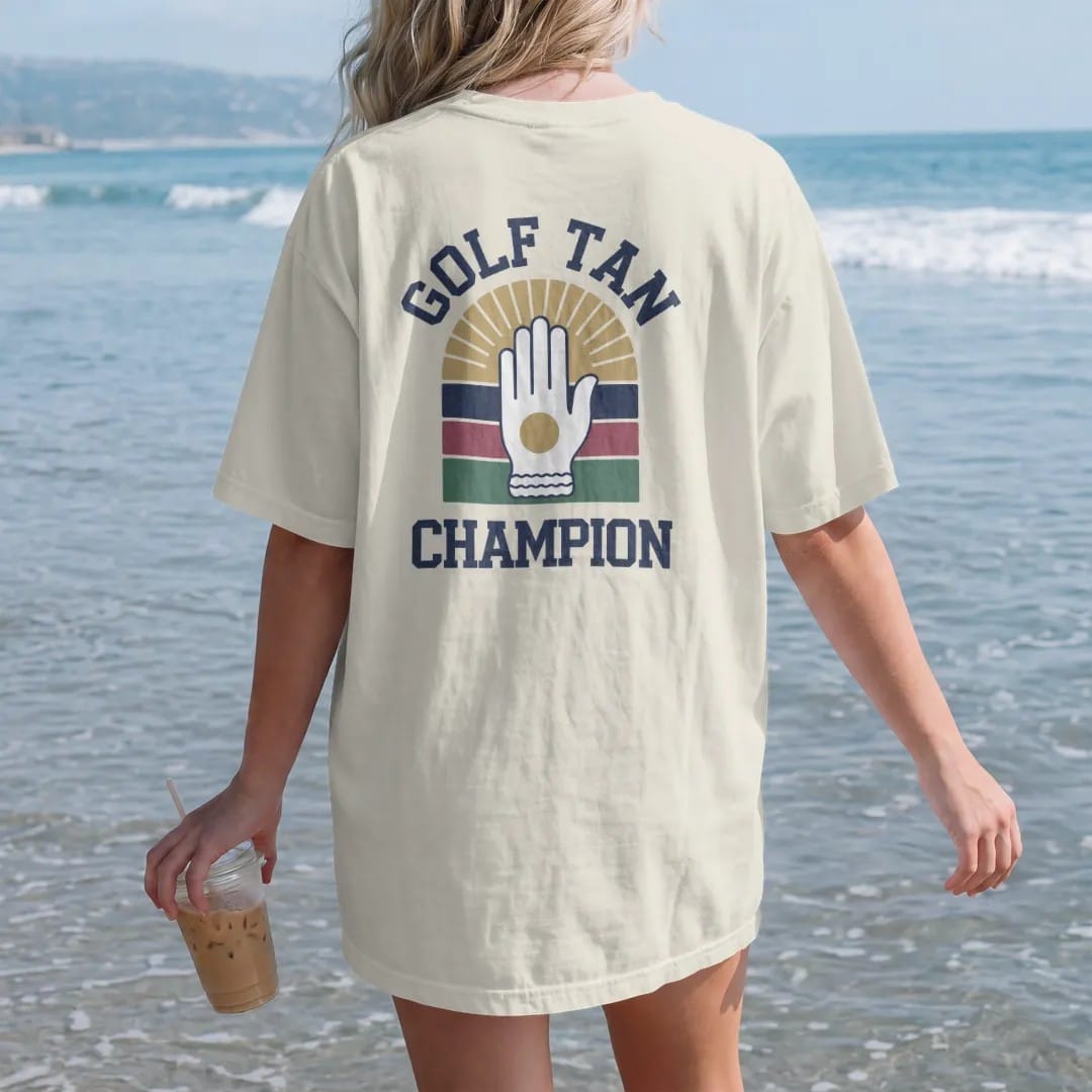 Woman on the beach in a Ivory tee, back view: a golfer's hand with a bright glove tan line under a big smiling sun, with "GOLF TAN" and "CHAMPION" in chunky faded collegiate block letters, printed in faded fairway green, washed navy, faded flag red…