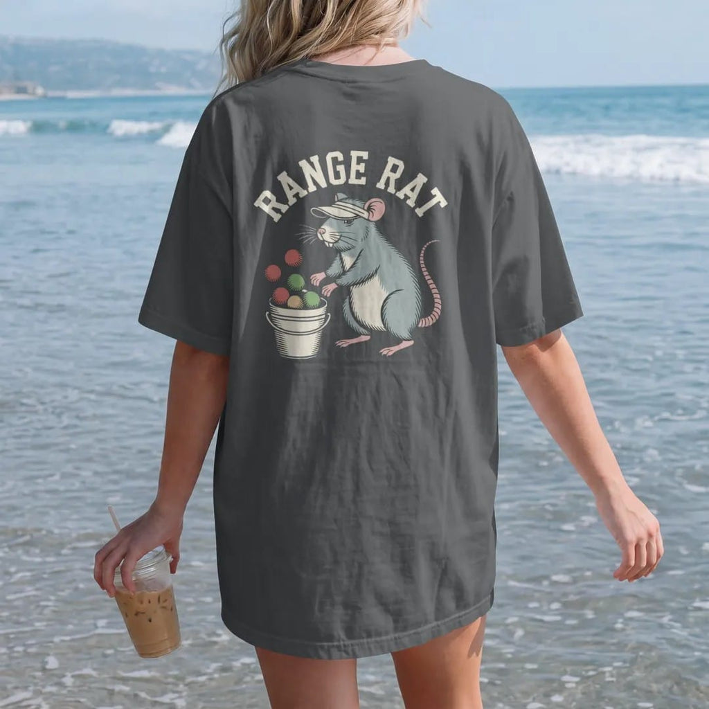 Woman on the beach in a Graphite tee, back view: a cheerful rat in a visor hitting balls from a bucket at a driving range, with "RANGE RAT" in loopy 1970s script, printed in faded clubhouse cream, sage grass green, washed flag red and sand gold…