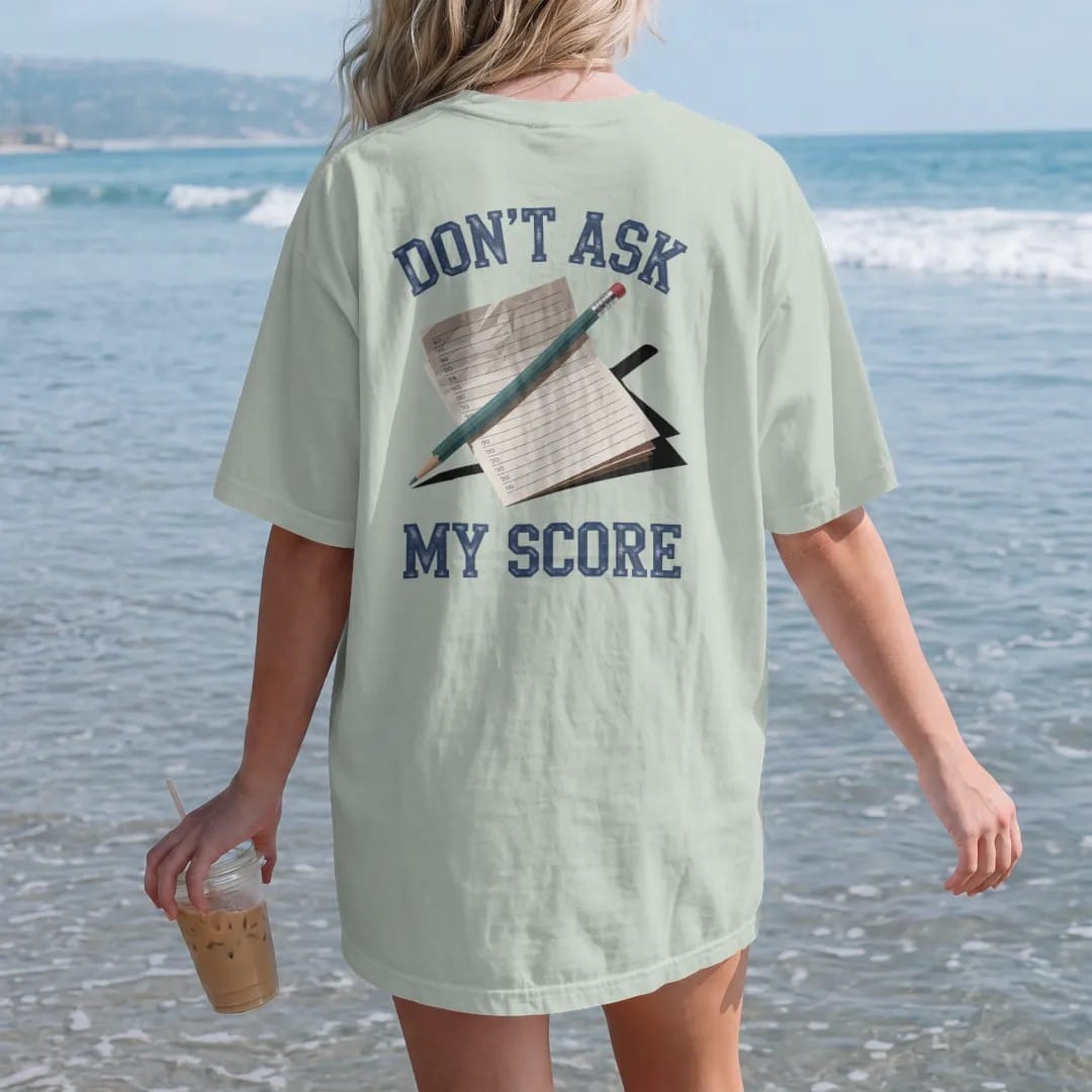 Woman on the beach in a Bay tee, back view: a stubby golf pencil on top of a crumpled scorecard, with "DON'T ASK" and "MY SCORE" in chunky faded collegiate block letters, printed in faded fairway green, washed navy, faded flag red and sand tan with…
