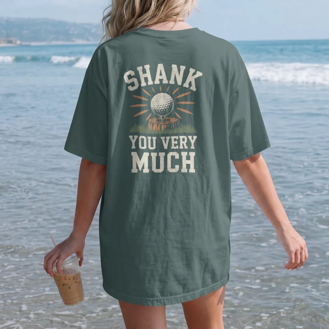 Woman on the beach in a Blue Spruce tee, back view: a golf ball ricocheting off a tree trunk with starbursts, with "SHANK" and "YOU VERY MUCH" in chunky faded collegiate block letters, printed in faded clubhouse cream, sage grass green, washed flag…