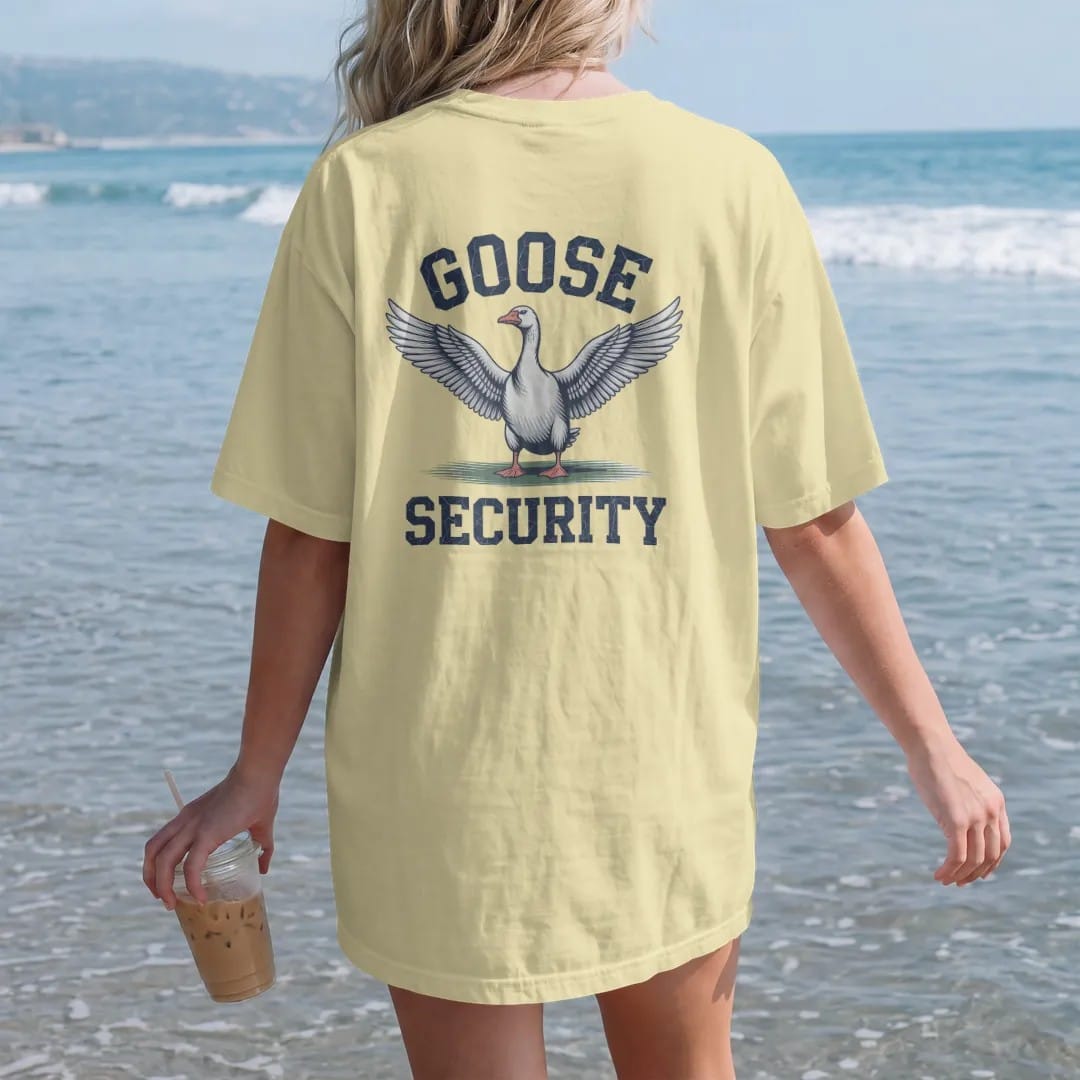 Woman on the beach in a Butter tee, back view: an angry goose with wings spread wide on a fairway, with "GOOSE" and "SECURITY" in chunky faded collegiate block letters, printed in faded fairway green, washed navy, faded flag red and sand tan with a…