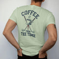 Man wearing a Light Green tee, seen from the back: a steaming coffee cup balanced on a golf tee with clubs behind, with "COFFEE" and "THEN TEE TIME" in loopy 1970s script, printed in faded fairway green, washed navy, faded flag red and sand tan…