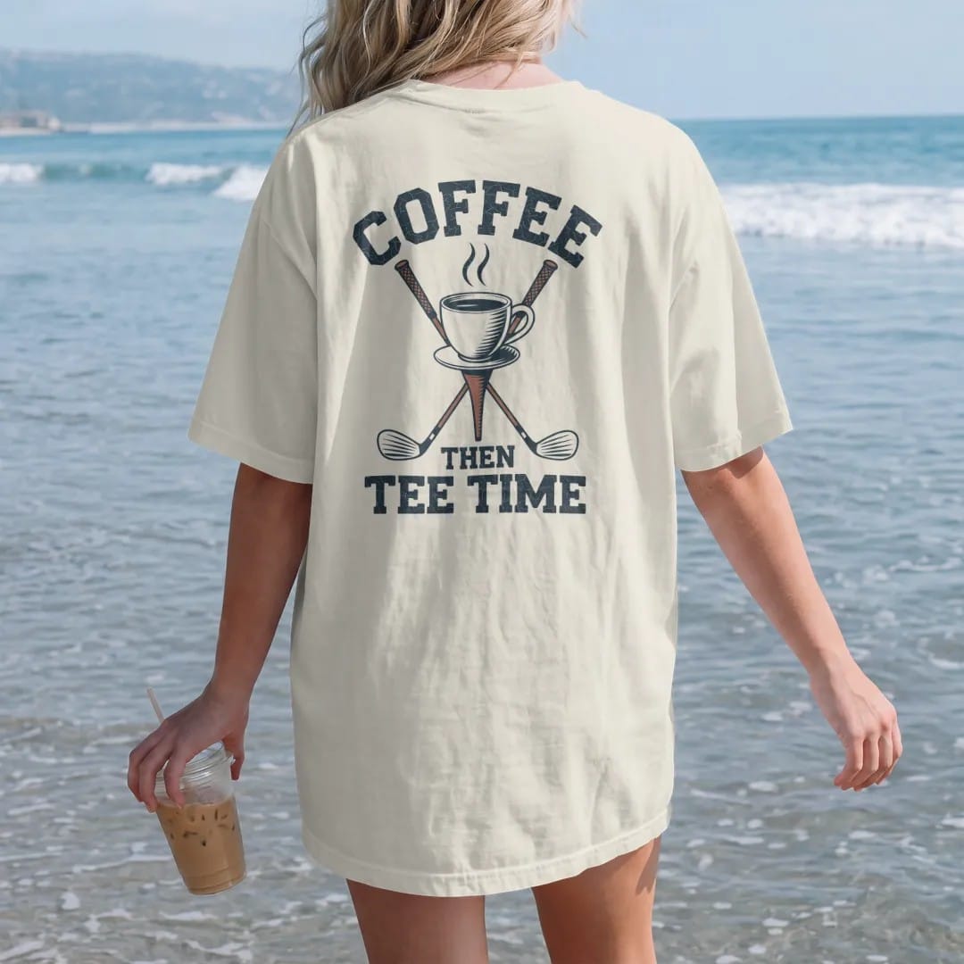 Woman on the beach in a Ivory tee, back view: a steaming coffee cup balanced on a golf tee with clubs behind, with "COFFEE" and "THEN TEE TIME" in loopy 1970s script, printed in faded fairway green, washed navy, faded flag red and sand tan with a…