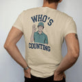 Man wearing a Sandstone tee, seen from the back: a golfer counting strokes on his fingers with a confused face, with "WHO'S" and "COUNTING" in chunky rounded 70s serif letters, printed in faded fairway green, washed navy, faded flag red and sand…