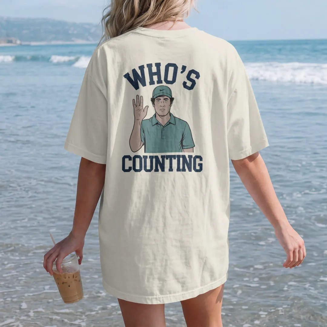 Woman on the beach in a Ivory tee, back view: a golfer counting strokes on his fingers with a confused face, with "WHO'S" and "COUNTING" in chunky rounded 70s serif letters, printed in faded fairway green, washed navy, faded flag red and sand tan…