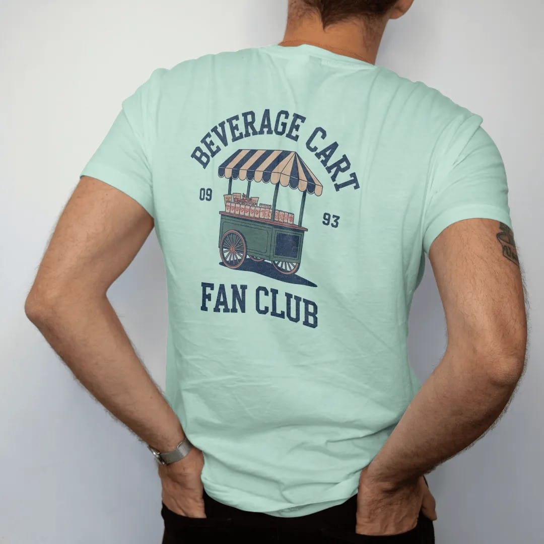 Man wearing a Chalky Mint tee, seen from the back: a vintage beverage cart with a striped awning loaded with cold cans and snacks, with "BEVERAGE CART" and "FAN CLUB" in loopy 1970s script, printed in faded fairway green, washed navy, faded flag…