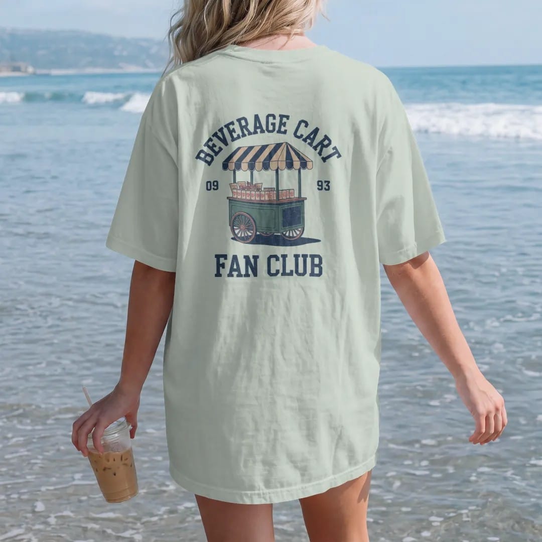 Woman on the beach in a Bay tee, back view: a vintage beverage cart with a striped awning loaded with cold cans and snacks, with "BEVERAGE CART" and "FAN CLUB" in loopy 1970s script, printed in faded fairway green, washed navy, faded flag red and…