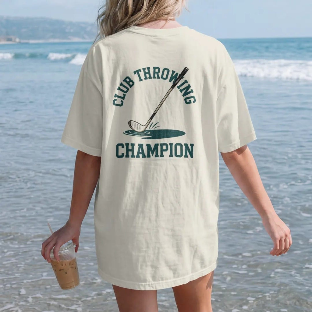 Woman on the beach in a Ivory tee, back view: a golf club tumbling end over end through the air toward a pond, with "CLUB THROWING" and "CHAMPION" in chunky faded collegiate block letters, printed in faded clubhouse cream, sage grass green, washed…