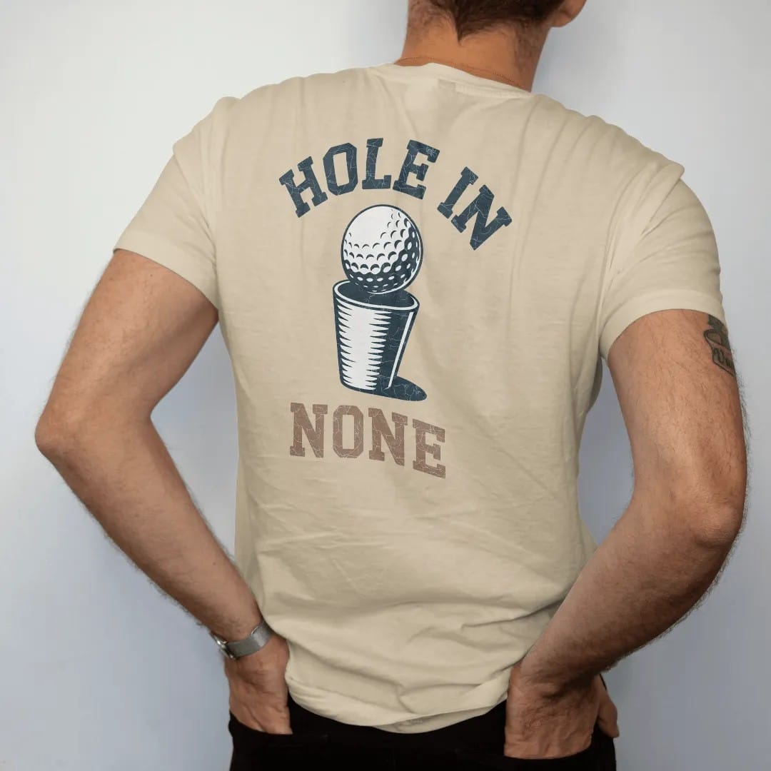 Man wearing a Sandstone tee, seen from the back: a golf ball lipping out of the cup with a dramatic spin, with "HOLE IN" and "NONE" in chunky rounded 70s serif letters, printed in faded fairway green, washed navy, faded flag red and sand tan with a…