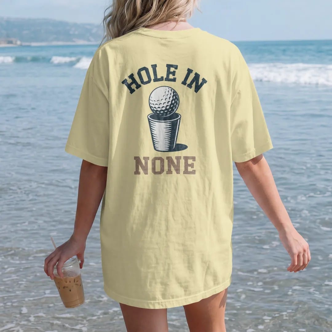 Woman on the beach in a Butter tee, back view: a golf ball lipping out of the cup with a dramatic spin, with "HOLE IN" and "NONE" in chunky rounded 70s serif letters, printed in faded fairway green, washed navy, faded flag red and sand tan with a…