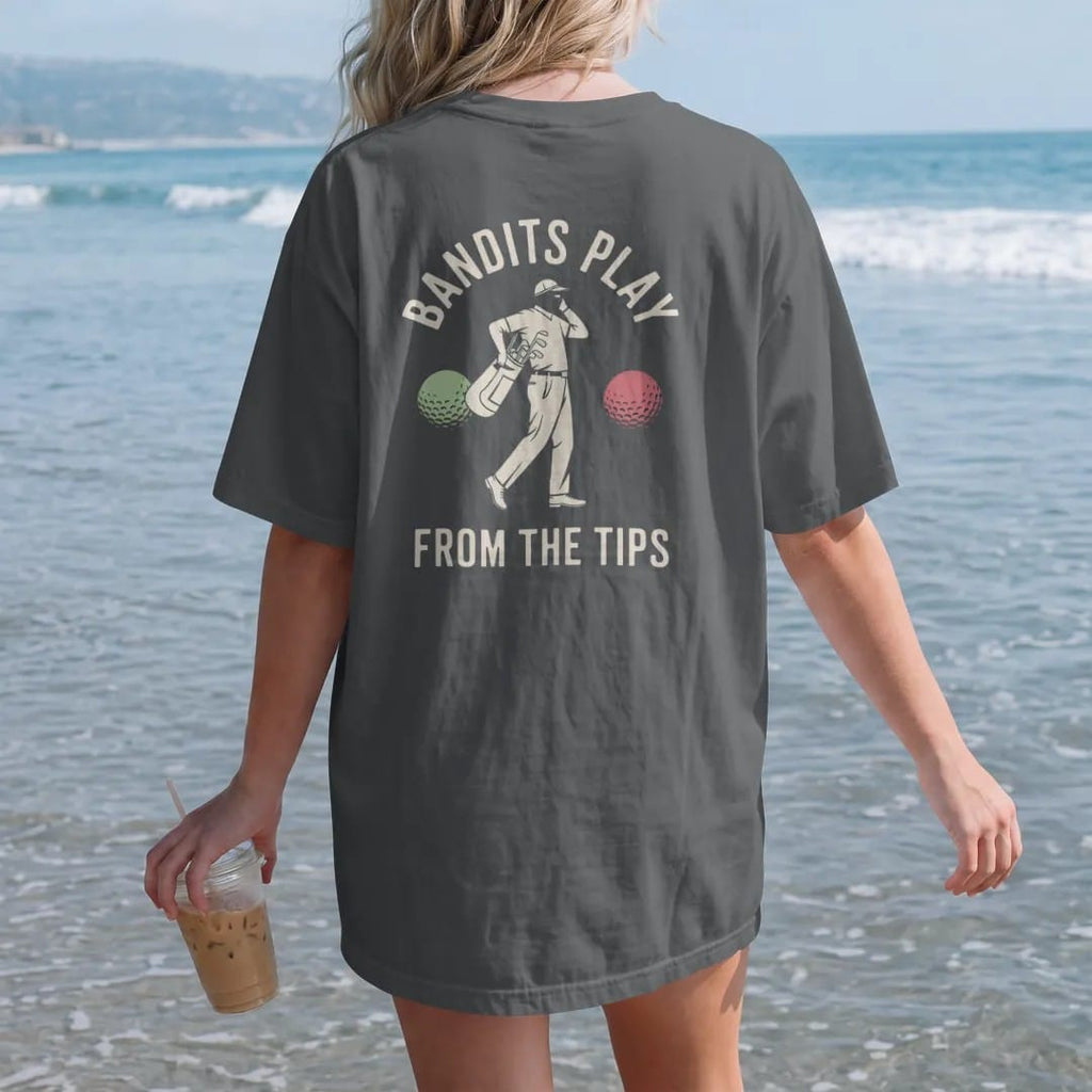 Woman on the beach in a Graphite tee, back view: a masked bandit golfer tiptoeing off with a bag of golf balls, with "BANDITS PLAY" and "FROM THE TIPS" in loopy 1970s script, printed in faded clubhouse cream, sage grass green, washed flag red and…