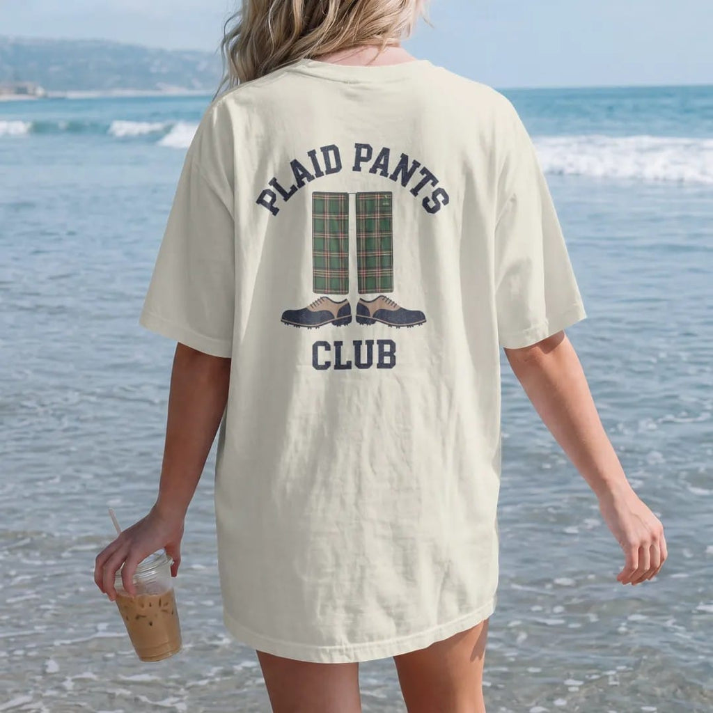 Woman on the beach in a Ivory tee, back view: a pair of loud plaid golf trousers above two-tone golf shoes, with "PLAID PANTS" and "CLUB" in loopy 1970s script, printed in faded fairway green, washed navy, faded flag red and sand tan with a…