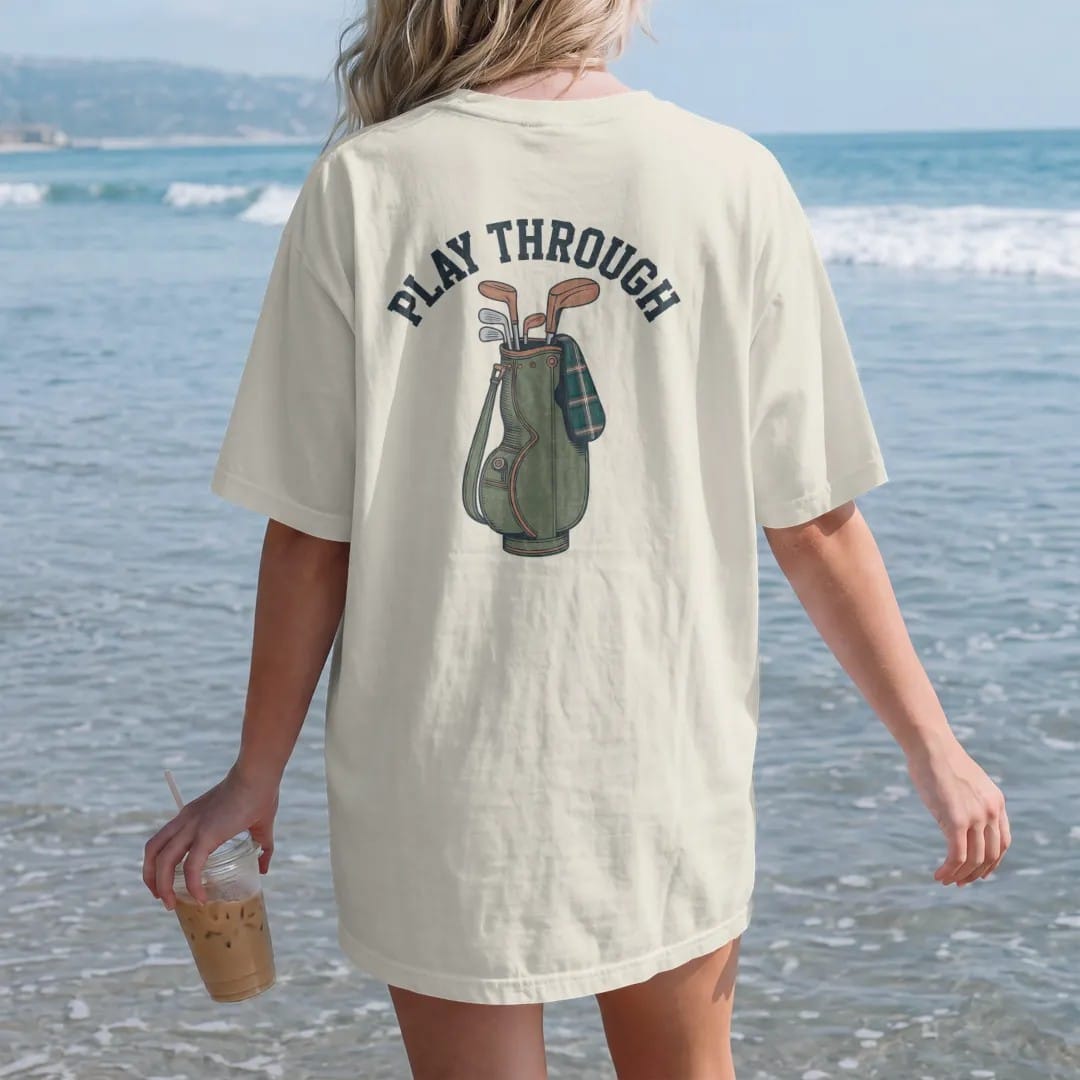 Woman on the beach in a Ivory tee, back view: a vintage leather golf bag with wooden-headed clubs and a tartan headcover, with "PLAY THROUGH" in chunky rounded 70s serif letters, printed in faded fairway green, washed navy, faded flag red and sand…