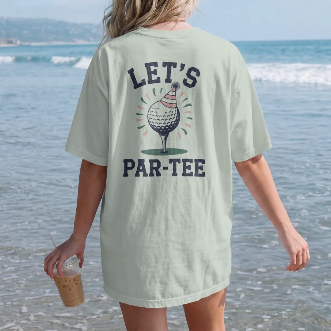 Woman on the beach in a Bay tee, back view: a golf ball on a tee wearing a party hat with confetti flying, with "LET'S" and "PAR-TEE" in loopy 1970s script, printed in faded fairway green, washed navy, faded flag red and sand tan with a cracked,…