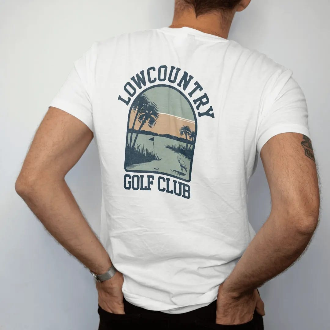 Man wearing a White tee, seen from the back: a golf hole beside a tidal marsh with palmetto trees and a wading heron, with "LOWCOUNTRY" and "GOLF CLUB" in chunky rounded 70s serif letters, printed in faded fairway green, washed navy, faded flag red…