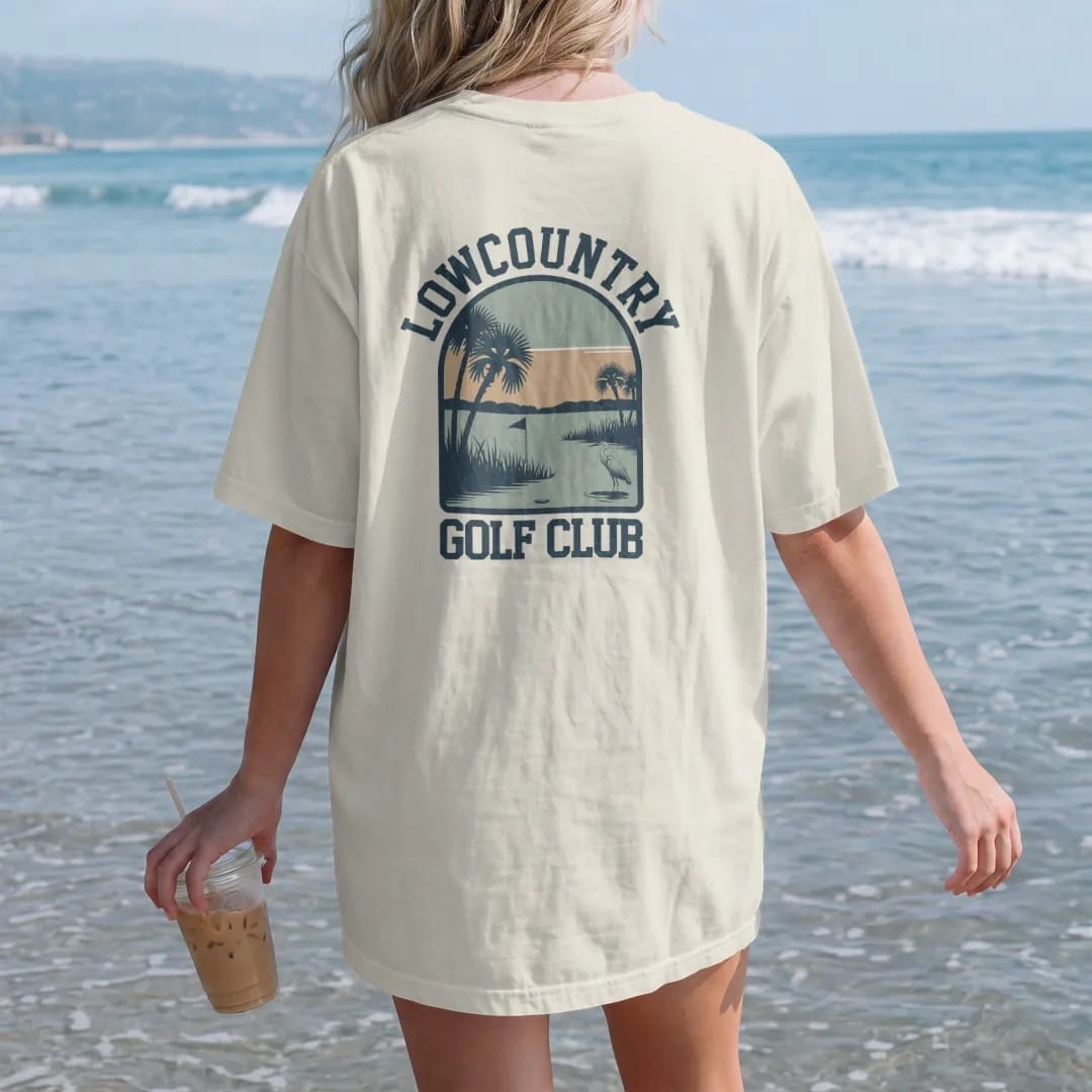 Woman on the beach in a Ivory tee, back view: a golf hole beside a tidal marsh with palmetto trees and a wading heron, with "LOWCOUNTRY" and "GOLF CLUB" in chunky rounded 70s serif letters, printed in faded fairway green, washed navy, faded flag…