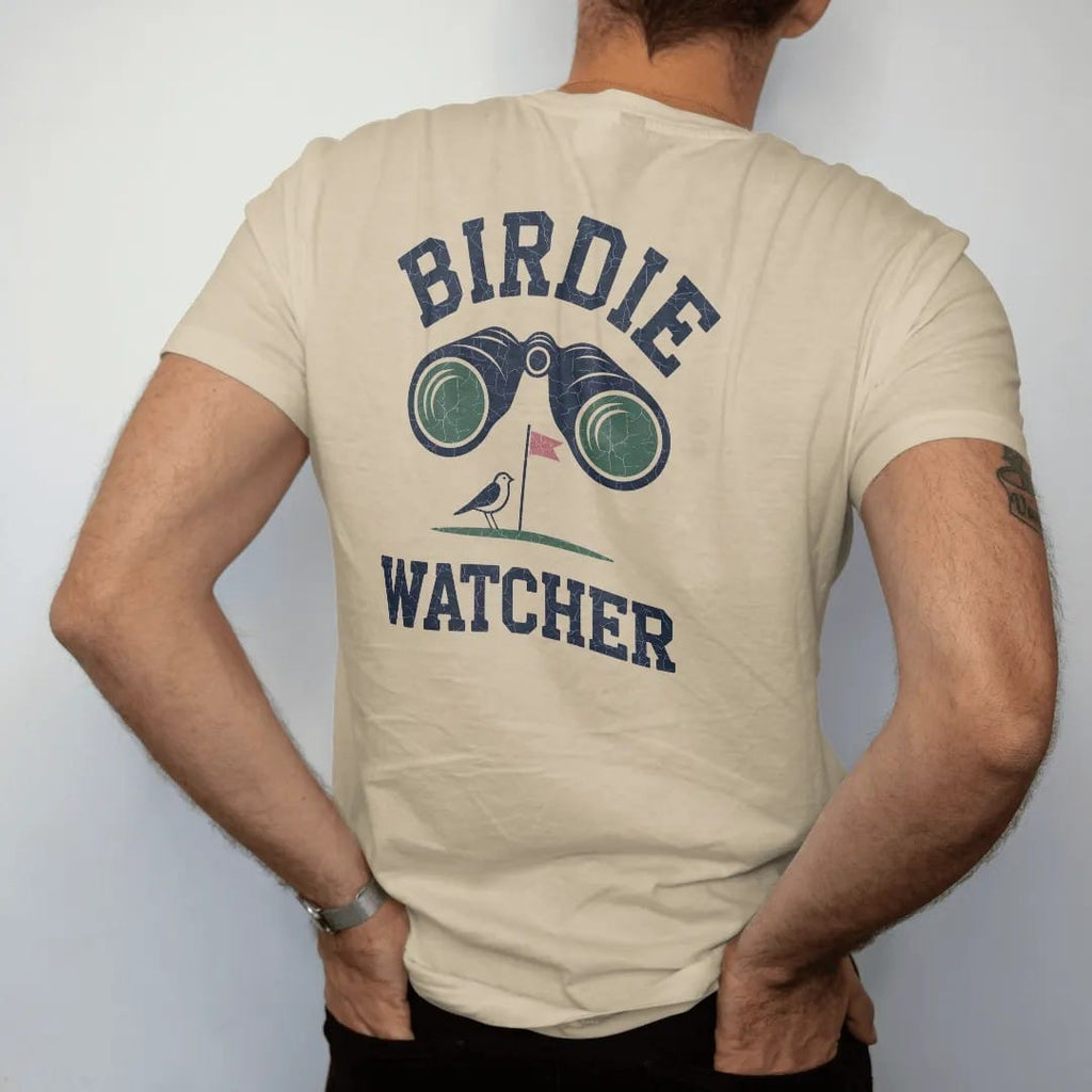 Man wearing a Sandstone tee, seen from the back: a pair of binoculars aimed at a tiny bird perched on a flagstick, with "BIRDIE" and "WATCHER" in chunky rounded 70s serif letters, printed in faded fairway green, washed navy, faded flag red and sand…