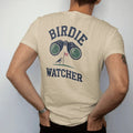 Man wearing a Sandstone tee, seen from the back: a pair of binoculars aimed at a tiny bird perched on a flagstick, with "BIRDIE" and "WATCHER" in chunky rounded 70s serif letters, printed in faded fairway green, washed navy, faded flag red and sand…
