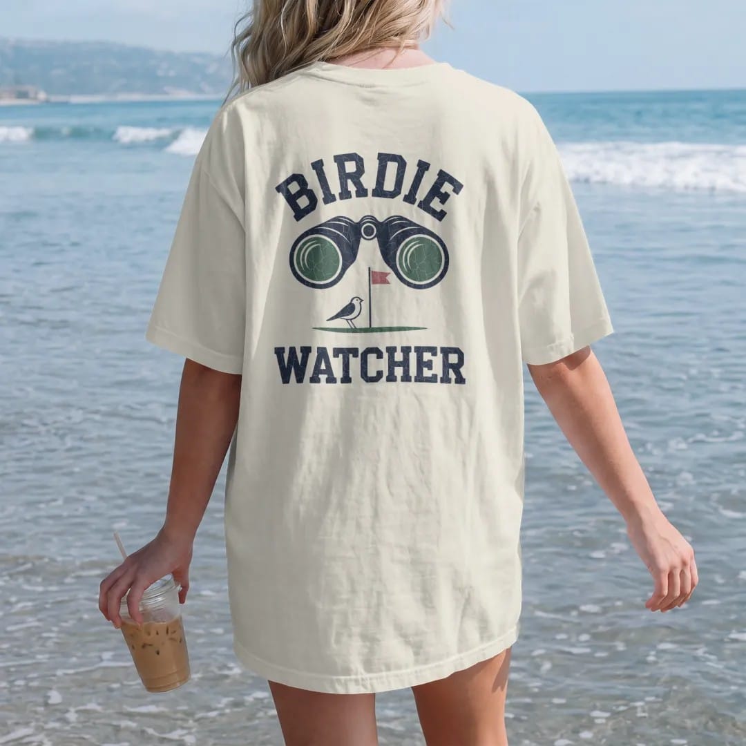 Woman on the beach in a Ivory tee, back view: a pair of binoculars aimed at a tiny bird perched on a flagstick, with "BIRDIE" and "WATCHER" in chunky rounded 70s serif letters, printed in faded fairway green, washed navy, faded flag red and sand…