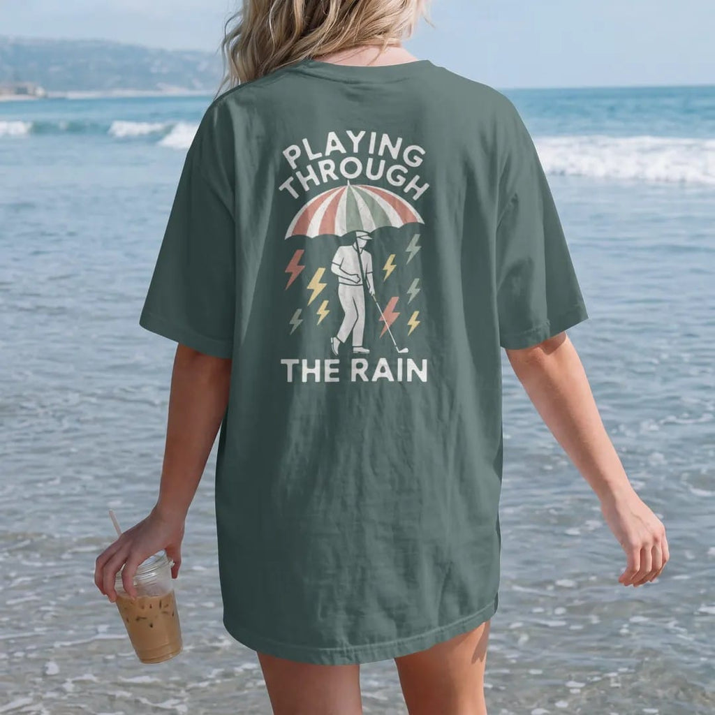 Woman on the beach in a Blue Spruce tee, back view: a golfer under a big striped umbrella with rain and lightning bolts, with "PLAYING THROUGH" and "THE RAIN" in loopy 1970s script, printed in faded clubhouse cream, sage grass green, washed flag…