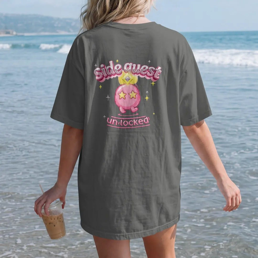 Woman on the beach in a Pepper tee, back view: blip the pixel slime under a yellow quest marker, with side quest arched in pink and silver chrome bubble letters and unlocked in a lime pixel banner.