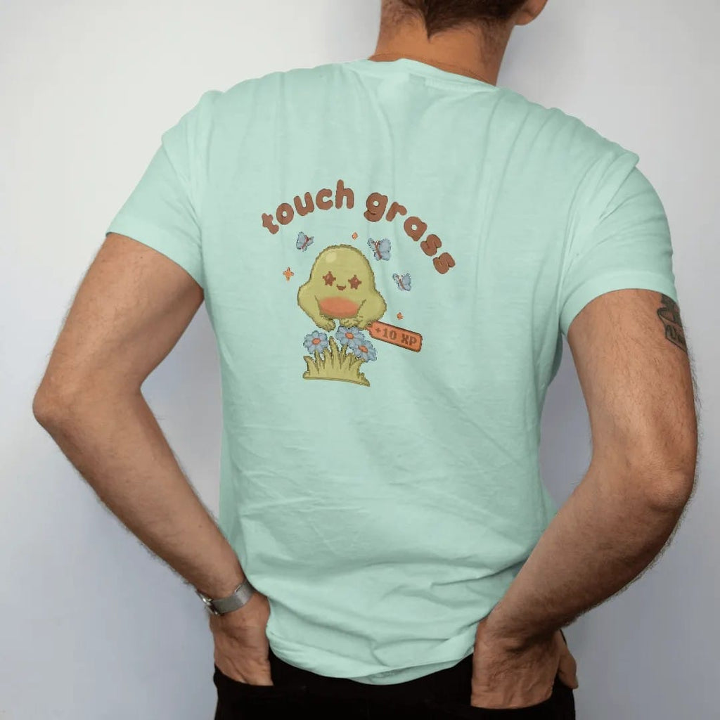 Man wearing a Chalky Mint tee, seen from the back: blip the pixel slime patting a tuft of grass and daisies under touch grass in rounded letters, with a +10 XP pixel popup, in moss green, rust and brown.