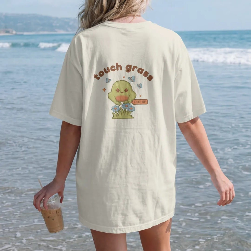 Woman on the beach in a Ivory tee, back view: blip the pixel slime patting a tuft of grass and daisies under touch grass in rounded letters, with a +10 XP pixel popup, in moss green, rust and brown.