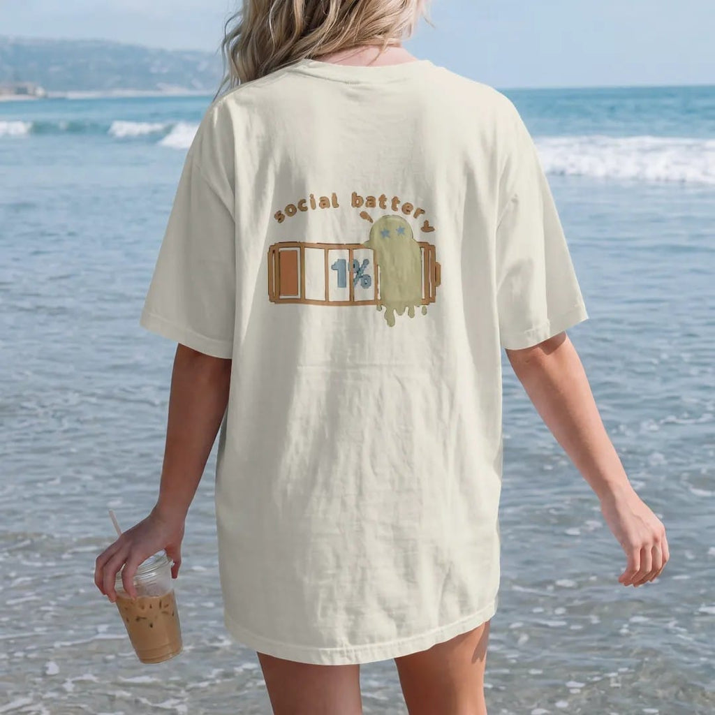 Woman on the beach in a Ivory tee, back view: blip the pixel slime melting against a nearly empty pixel battery reading 1%, under social battery in brown and rust rounded letters.
