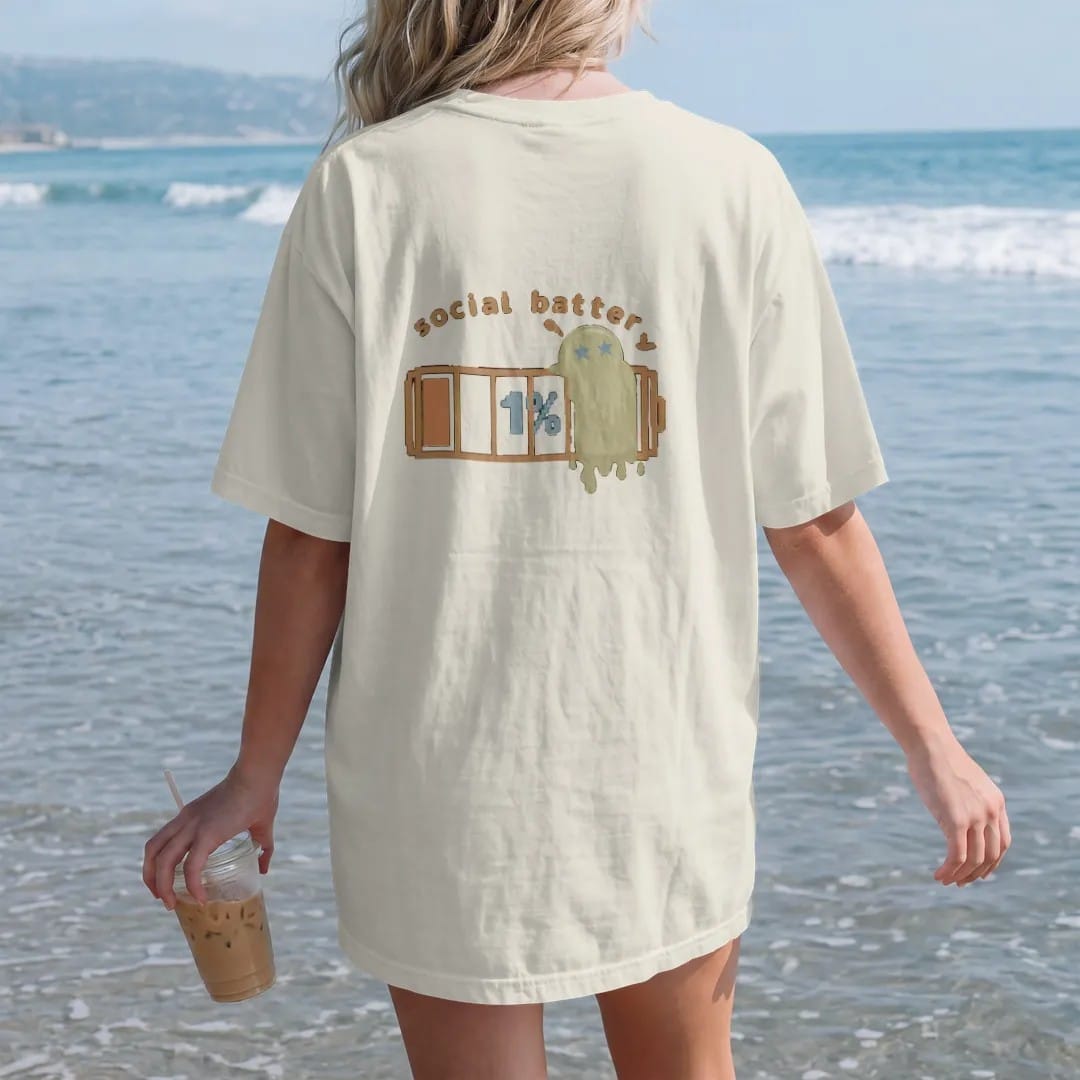 Woman on the beach in a Ivory tee, back view: blip the pixel slime melting against a nearly empty pixel battery reading 1%, under social battery in brown and rust rounded letters.