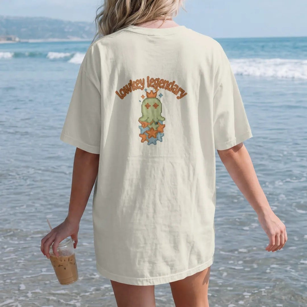 Woman on the beach in a Ivory tee, back view: blip the pixel slime wearing a tilted crown on a stack of rarity stars, under lowkey legendary in brown, rust and moss wavy letters.