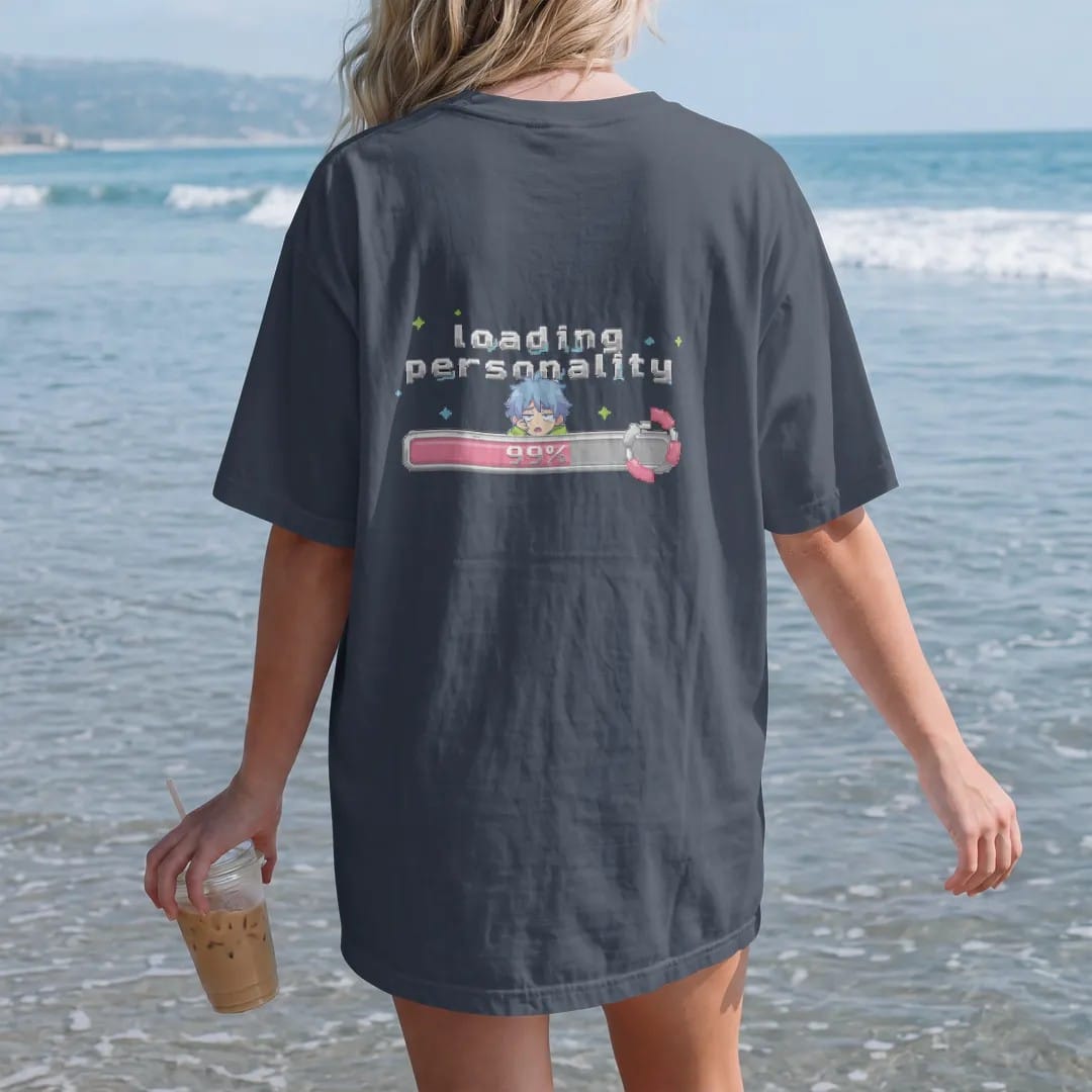 Woman on the beach in a Midnight tee, back view: a nearly full pixel loading bar reading 99% with a sleepy anime-style kid peeking over it, under loading personality in pink, lime and blue pixel letters.