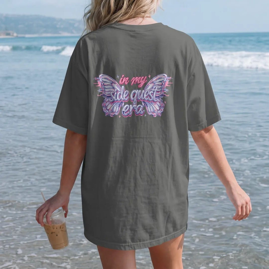 Woman on the beach in a Pepper tee, back view: two glossy chrome butterflies with glitch trails framing in my in brush script and side quest era in silver, lilac and pink chrome bubble letters.