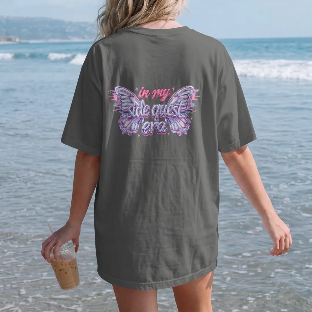 Woman on the beach in a Pepper tee, back view: two glossy chrome butterflies with glitch trails framing in my in brush script and side quest era in silver, lilac and pink chrome bubble letters.
