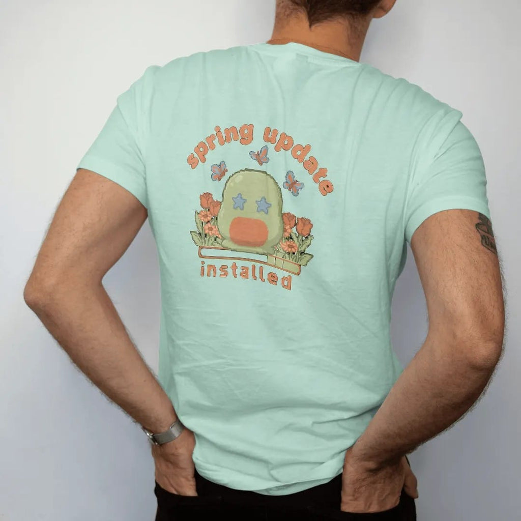 Man wearing a Chalky Mint tee, seen from the back: blip the pixel slime in a patch of pixel tulips and daisies with butterflies, under spring update in bubbly letters and installed in pixel type, in moss, rust, brown and denim.