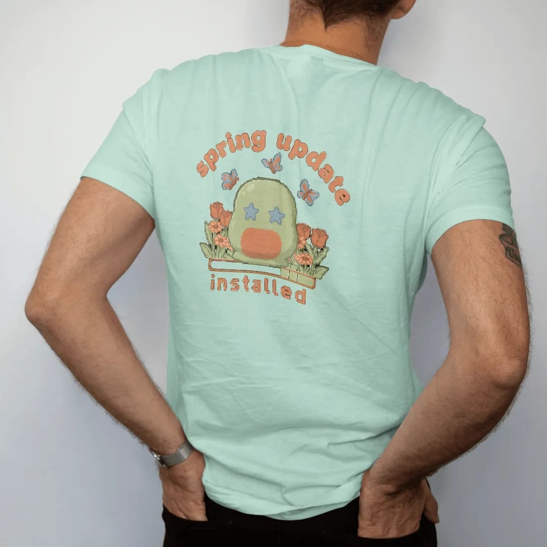 Man wearing a Chalky Mint tee, seen from the back: blip the pixel slime in a patch of pixel tulips and daisies with butterflies, under spring update in bubbly letters and installed in pixel type, in moss, rust, brown and denim.