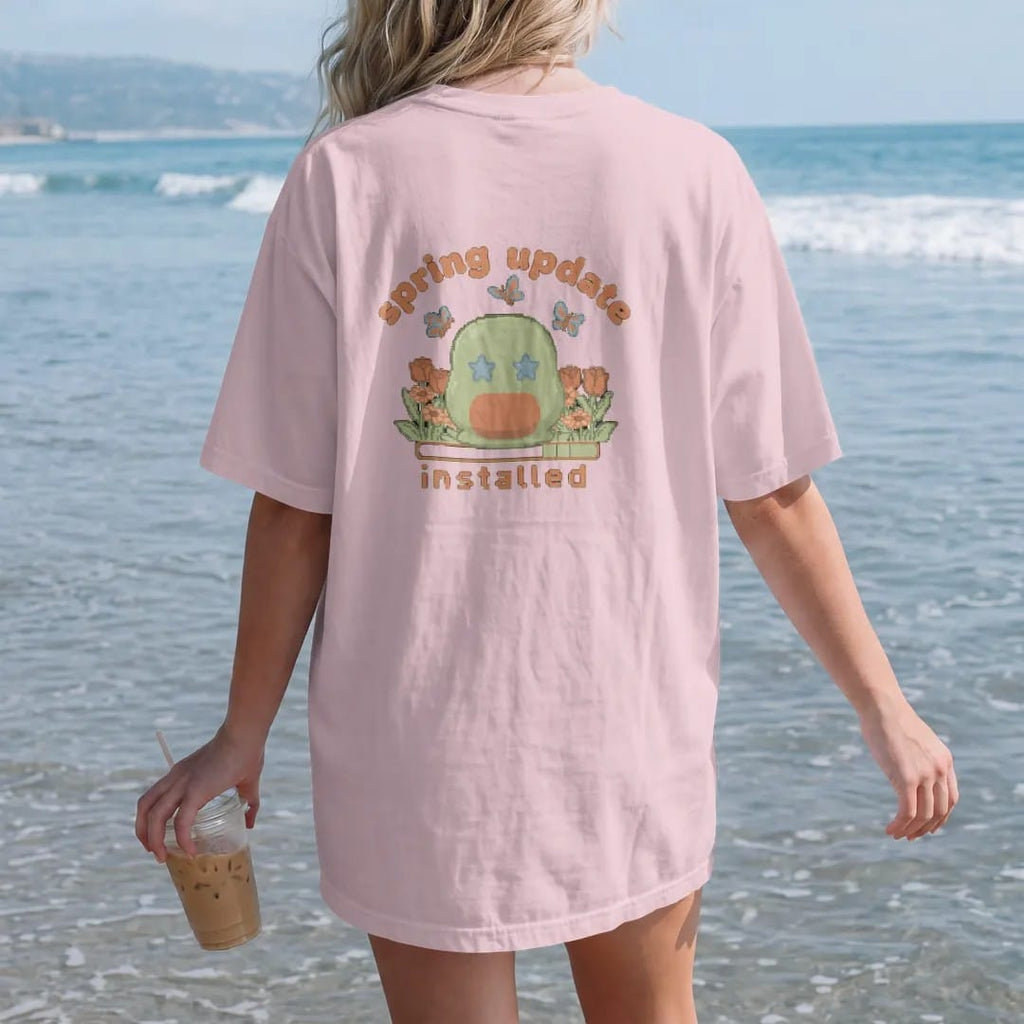 Woman on the beach in a Blossom tee, back view: blip the pixel slime in a patch of pixel tulips and daisies with butterflies, under spring update in bubbly letters and installed in pixel type, in moss, rust, brown and denim.
