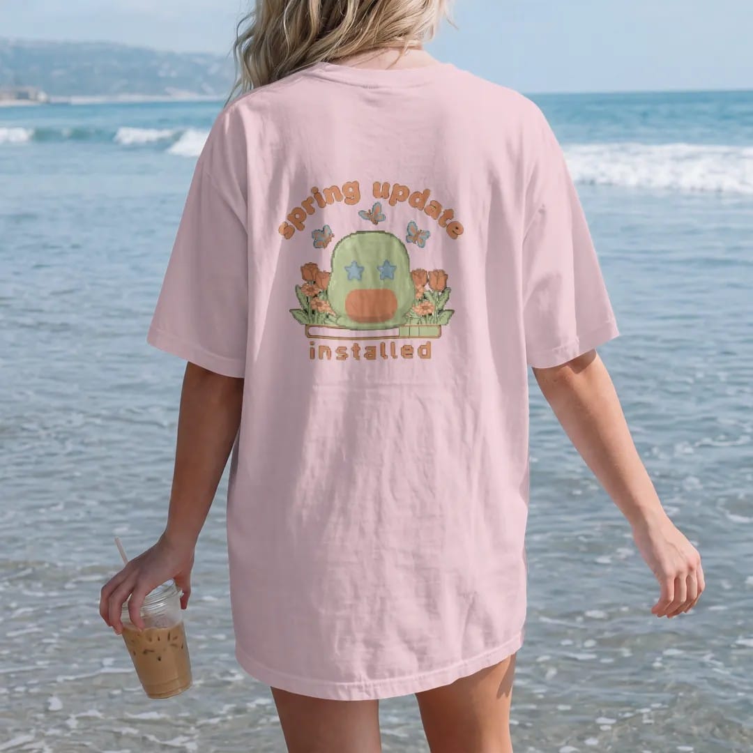 Woman on the beach in a Blossom tee, back view: blip the pixel slime in a patch of pixel tulips and daisies with butterflies, under spring update in bubbly letters and installed in pixel type, in moss, rust, brown and denim.