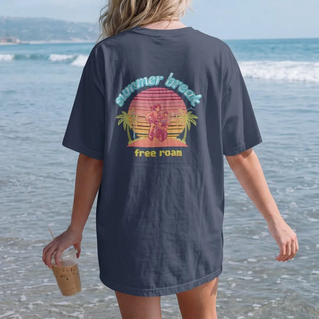Woman on the beach in a Navy tee, back view: two anime-style teens biking toward a striped pixel sunset with neon palm trees, under summer break in chrome bubble letters and free roam in pixel type, in pink, orange, lime and blue.