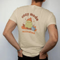 Man wearing a Sandstone tee, seen from the back: blip the pixel slime in a knit beanie burrowed in autumn leaves beside a mug of cocoa, under cozy mode in rounded letters and activated in pixel type, in rust, brown and moss.