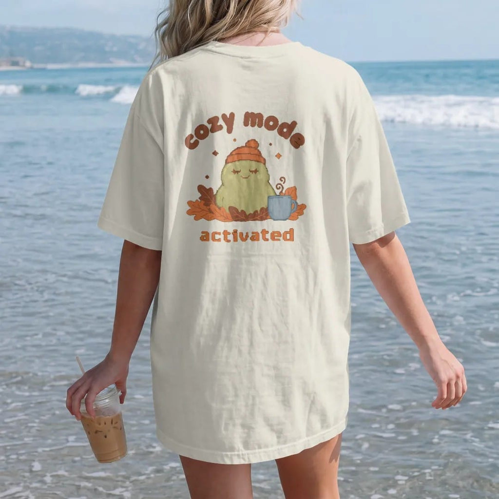 Woman on the beach in a Ivory tee, back view: blip the pixel slime in a knit beanie burrowed in autumn leaves beside a mug of cocoa, under cozy mode in rounded letters and activated in pixel type, in rust, brown and moss.