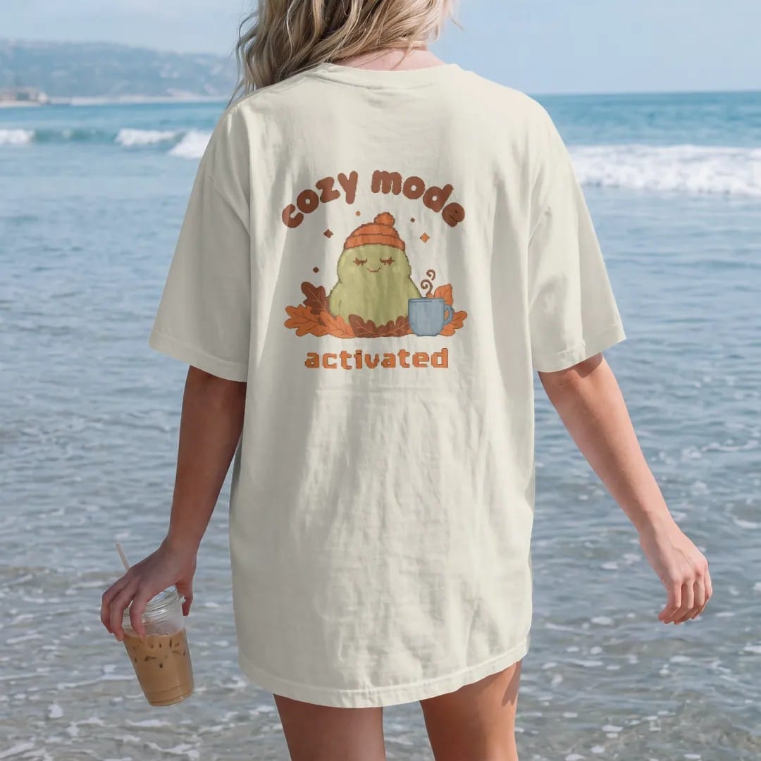 Woman on the beach in a Ivory tee, back view: blip the pixel slime in a knit beanie burrowed in autumn leaves beside a mug of cocoa, under cozy mode in rounded letters and activated in pixel type, in rust, brown and moss.